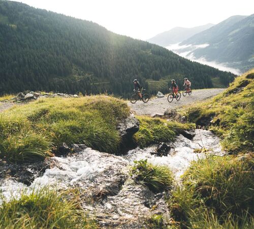 Drei Mountainbiker fahren auf einem breiten Bergweg entlang eines Gebirgsbachs mit Blick auf bewaldete Alpenlandschaft