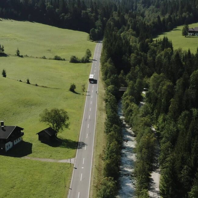 Bus fährt auf einer Landstraße umgeben von grünen Wiesen, Wald und Fluss.