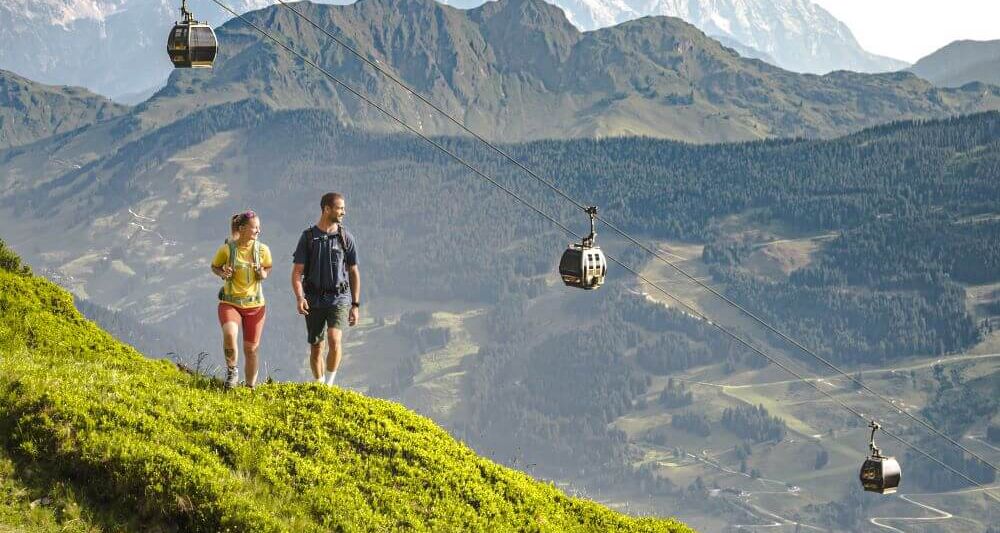 Über 2 Wanderern fährt die Sommerbergbahn, im Hintergrund 2 imposante Bergkämme.