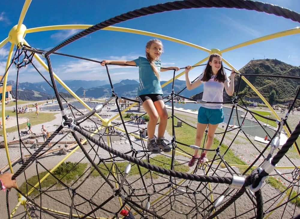 Kinder stehen im Kletternetz, im Hintergrund Sand  & Wasserspielplätze und Berge