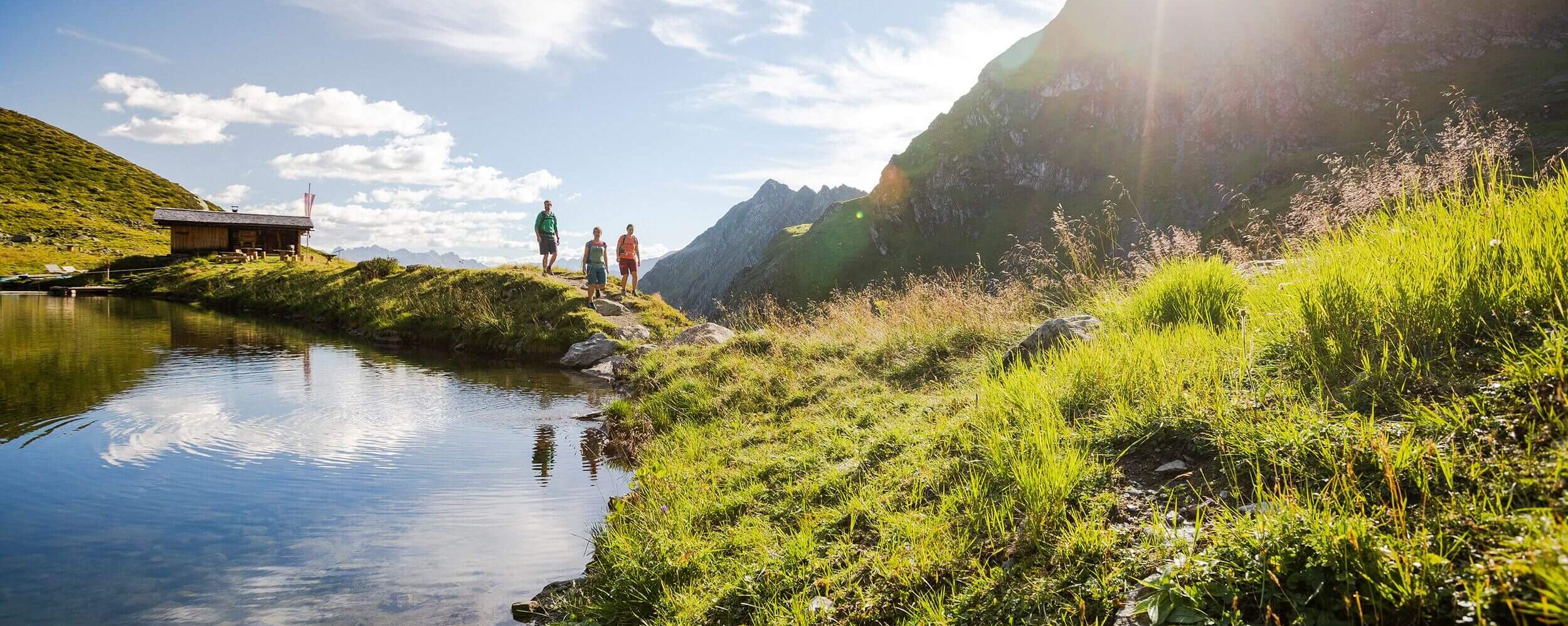 Familie beim Wandern entlang eines Bergsees mit traditioneller Tiroler Hütte und alpiner Bergkulisse im Hintergrund