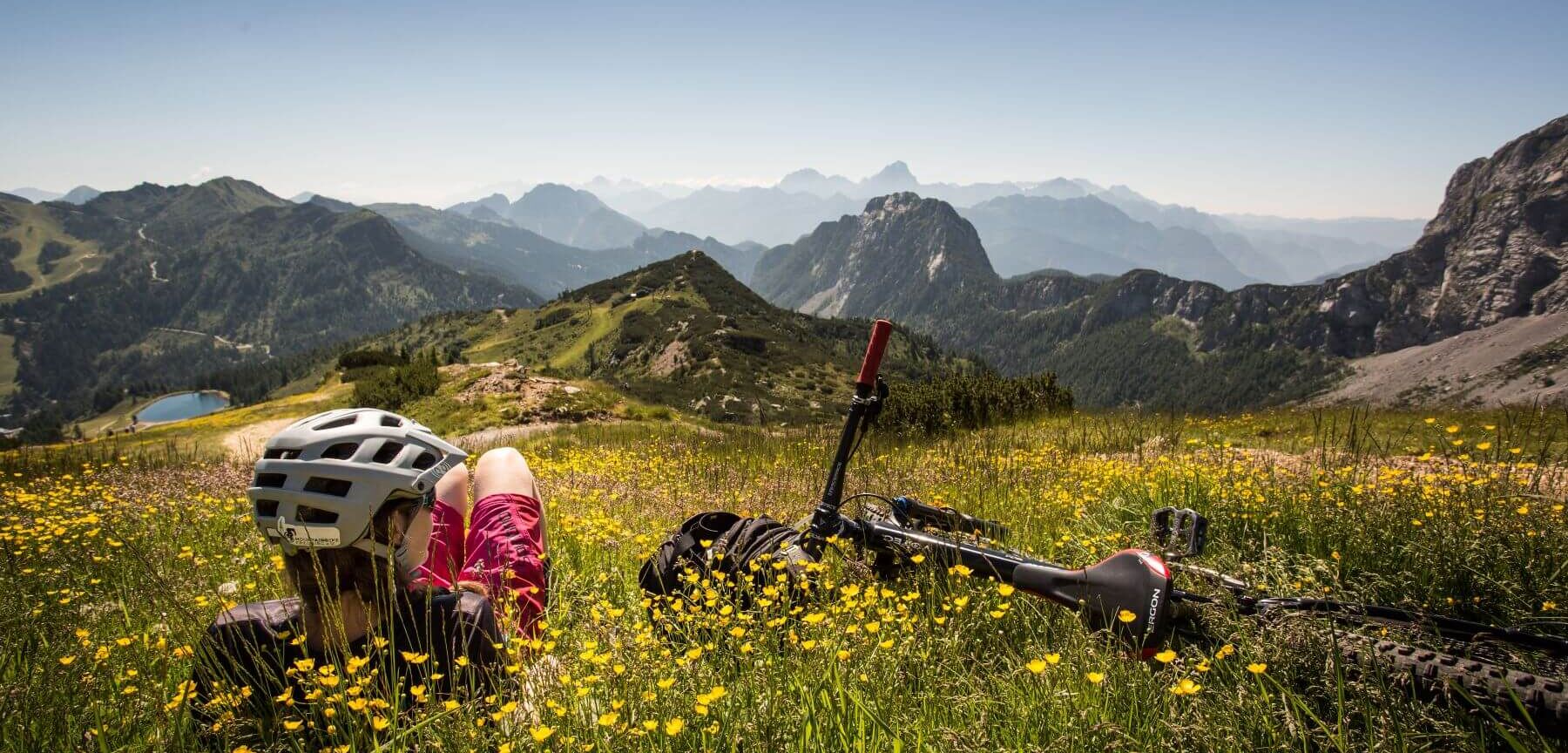 Bikerin entspannt neben ihrem Fahrrad auf einer blühenden Bergwiese und blickt auf eine beeindruckende Alpenlandschaft