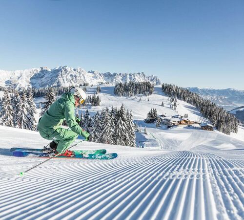 Eine Skifahrerin gleitet über die frisch präparierte Piste, umgeben von verschneiten Almen und einer majestätischen Bergkulisse