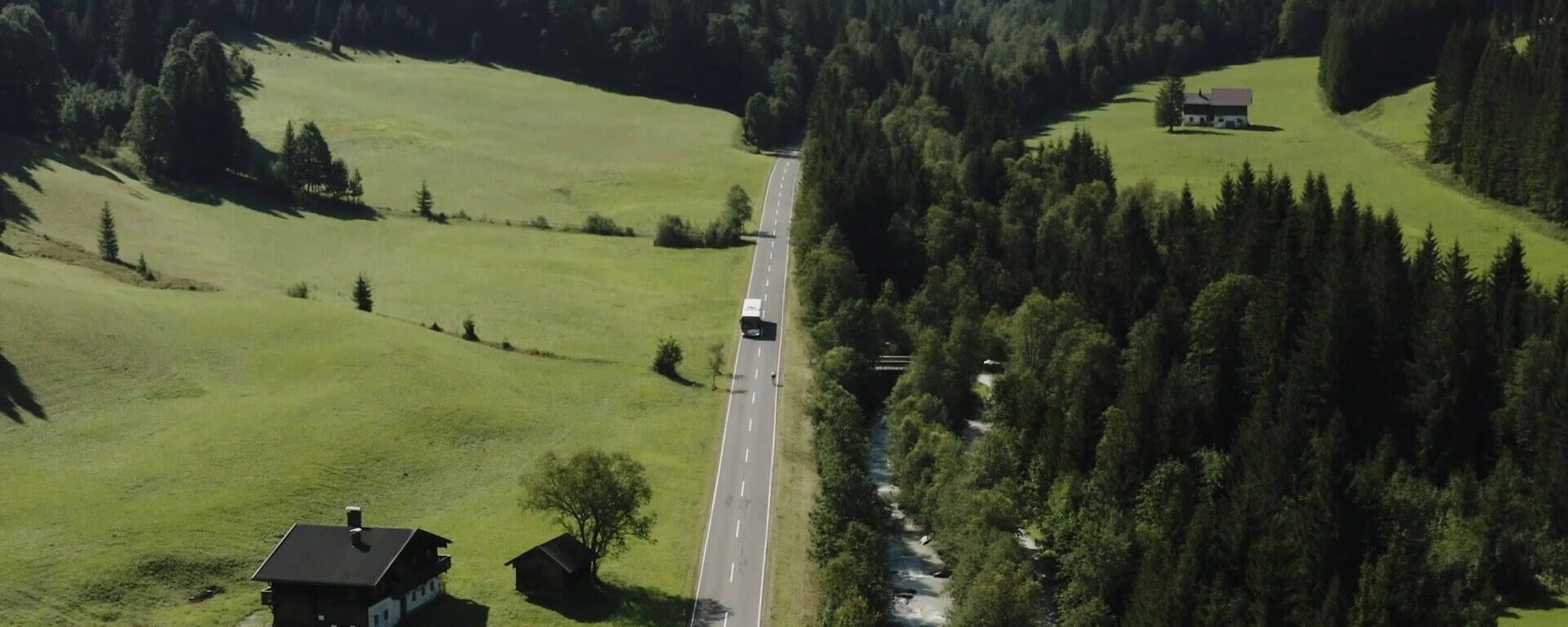 Bus travelling on a country road surrounded by green meadows, forest and river.