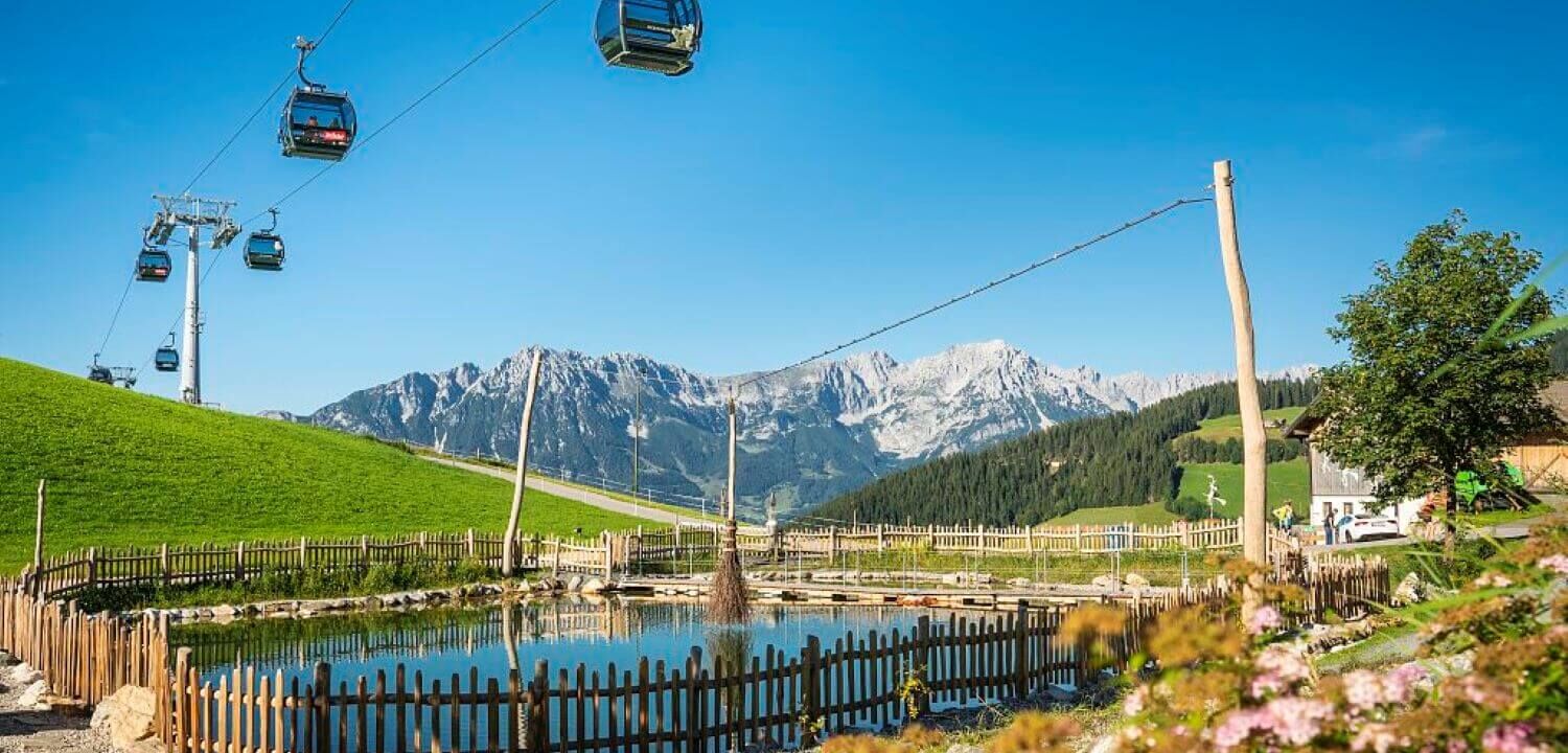 Sommerbergbahn schwebt über einen eingezäunten Teich, im Hintergrund das Bergpanorama