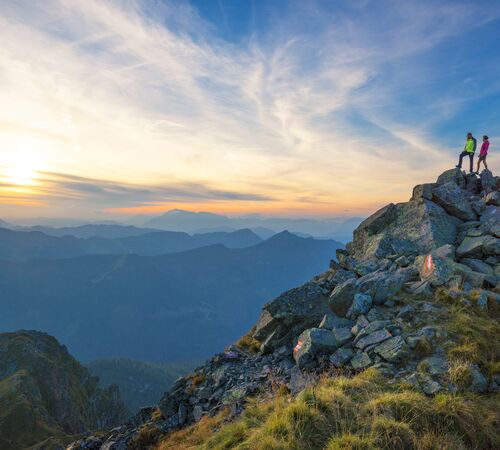 Zwei Wanderer stehen am Gipfelkreuz und blicken in die abendliche Berglandschaft mit weitem Panorama und warmem Licht