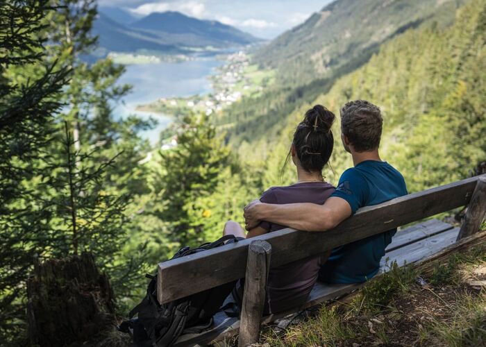 Ein Paar macht Rast auf einer Bank im Wald und genießt beim Wandern in Kärnten den Ausblick vom Berg auf einen See zwischen umliegenden Bergen