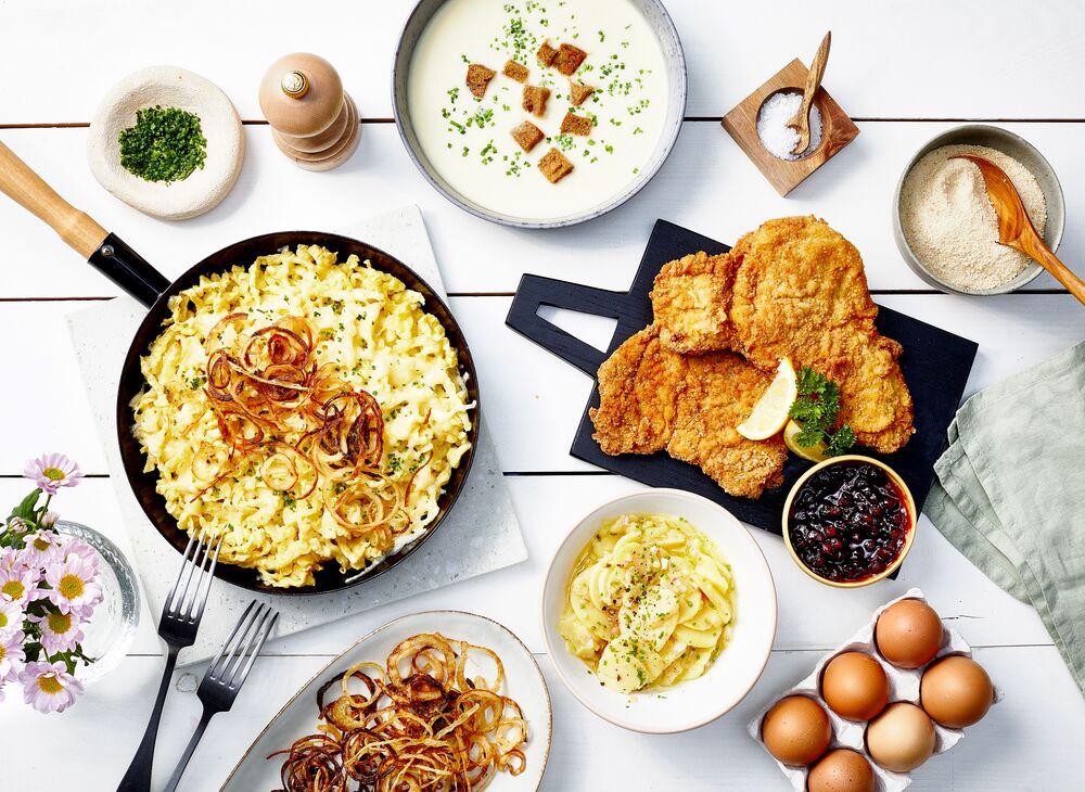 Auf einem Holztisch stehen regionale Spezialitäten wie goldbraune Käsespätzle mit Röstzwiebeln, ein knuspriges Schnitzel und frischer Kartoffelsalat.