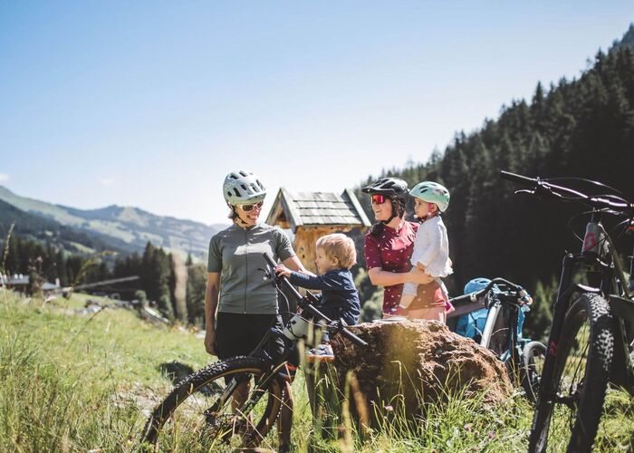 Zwei Mütter mit Fahrradhelmen und ihren Kindern machen Rast mit modernen E-Bikes auf einer Almwiese inmitten einer alpinen Berglandschaft.