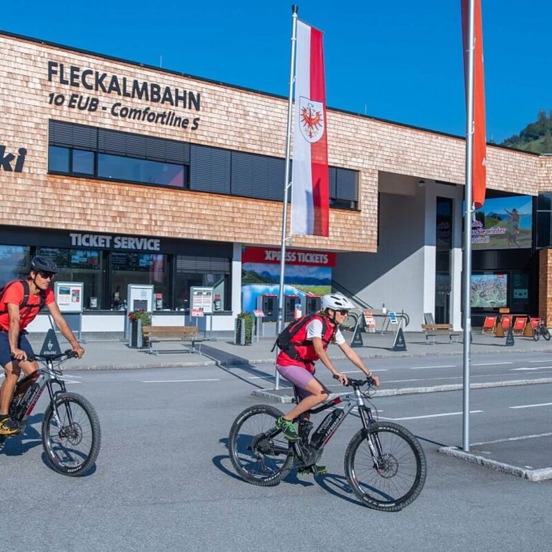2 E-Biker radeln zur Talstation einer Sommerbergbahn.