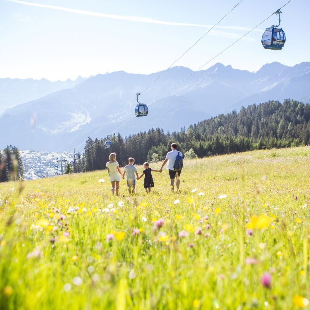 Eine Familie wandert über eine Bergwiese, oberhalb fährt eine Gondelbahn und bietet einfachen Zugang zum Wandergebiet