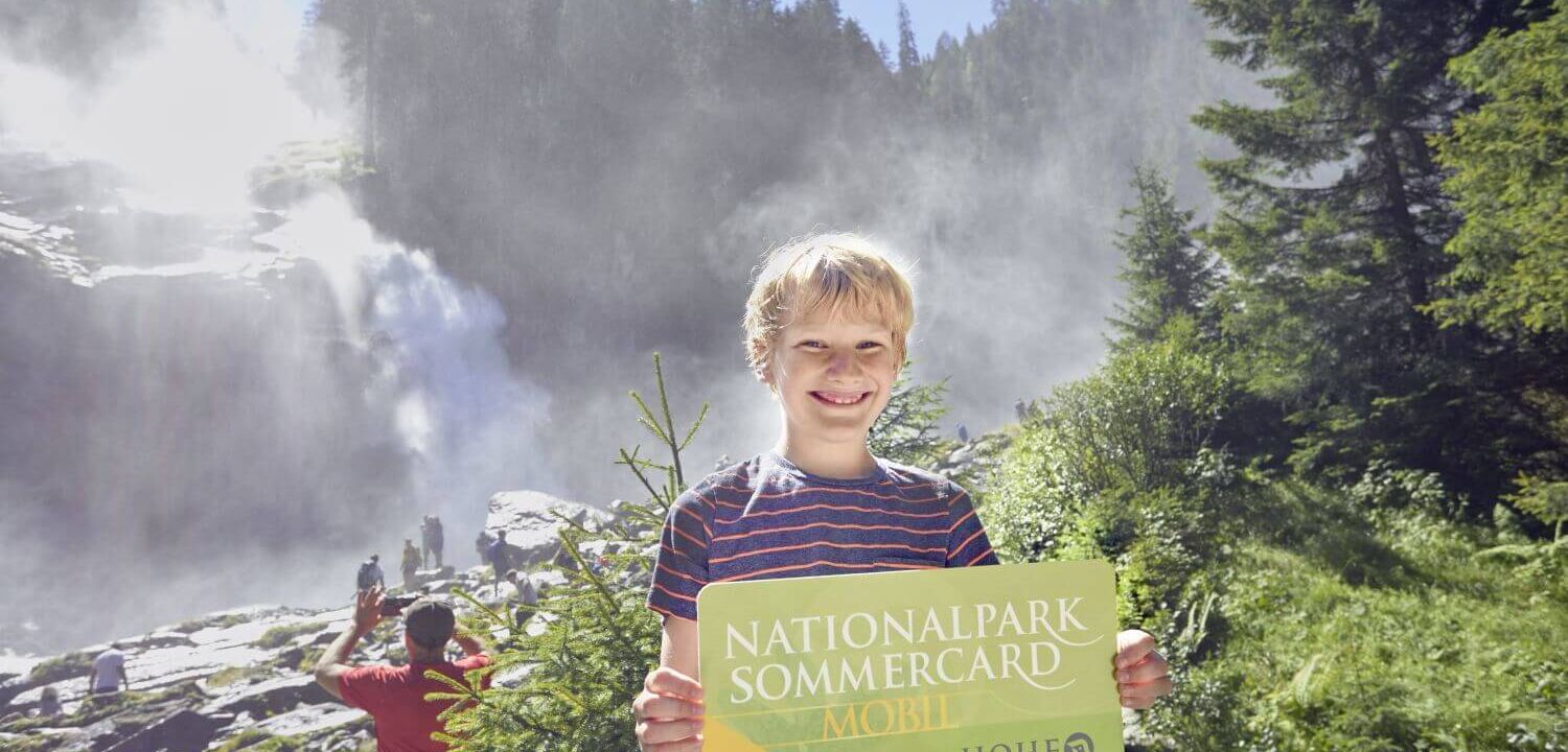 Junge steht mit der Nationalpark Sommercard in der Hand vor den Krimmler Wasserfällen.