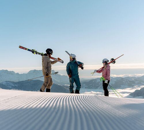 3 skier friends stand in the sun on the fresh piste above the sea of fog.