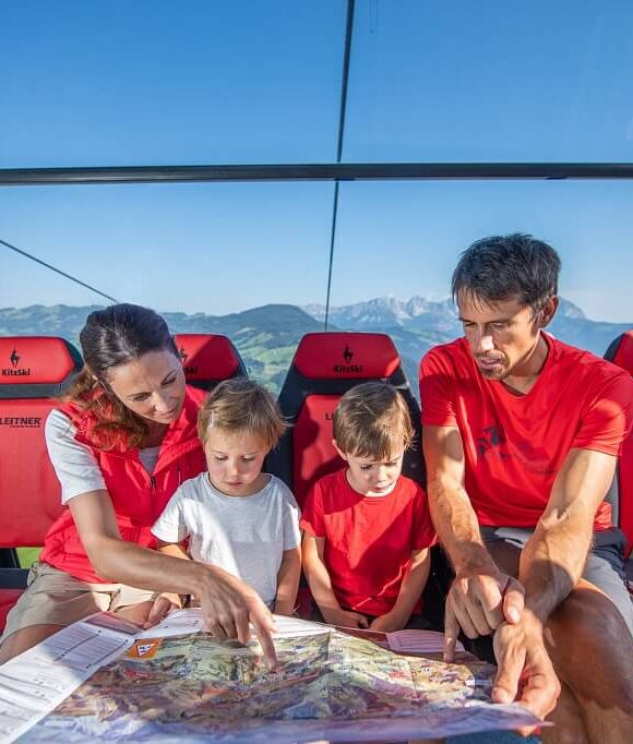 Familie mit 2 Kindern sitzt in einer verglasten Panoramagondel und studiert die Wanderkarte.