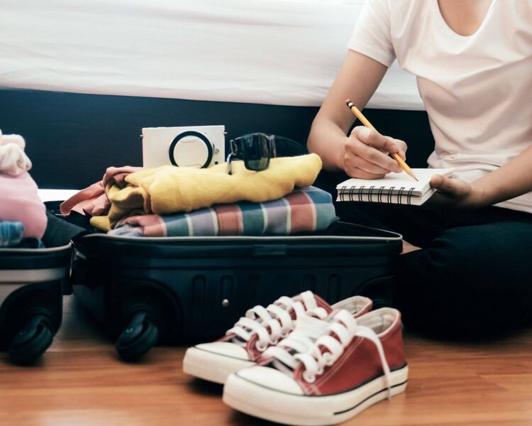 Junge Frau mit einer Checkliste sitzt vor ihrem offenen, vollbepackten Koffer auf dem Boden.