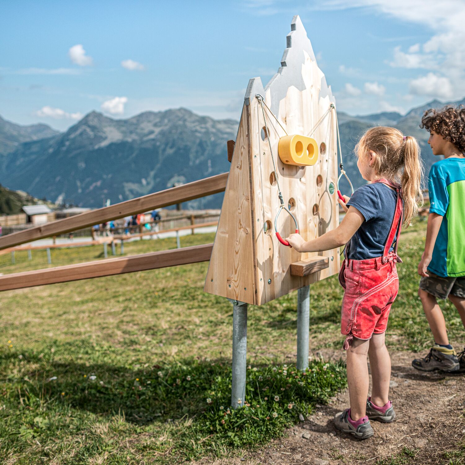 Zwei Kinder in Wanderkleidung spielen auf einer Alm bei einer Kugelbahn aus Holz, im Hintergrund ragen imposante Berge in den Himmel
