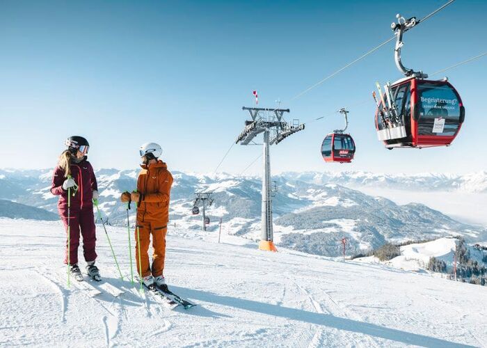 Zwei Skifahrer stehen auf der Piste unterhalb einer Kabinenbahn.