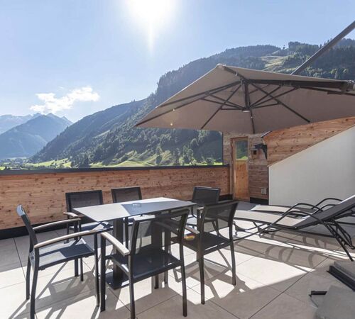 Terrasse einer Ferienwohnung mit Tisch, Stühlen, zwei Liegestühlen und Sonnenschirm – traumhafter Ausblick auf grüne Wälder und imposante Berggipfel im Salzburger Land.