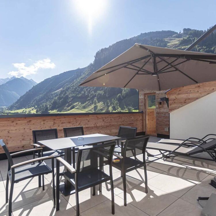 Terrasse einer Ferienwohnung mit Tisch, Stühlen, zwei Liegestühlen und Sonnenschirm – traumhafter Ausblick auf grüne Wälder und imposante Berggipfel im Salzburger Land.