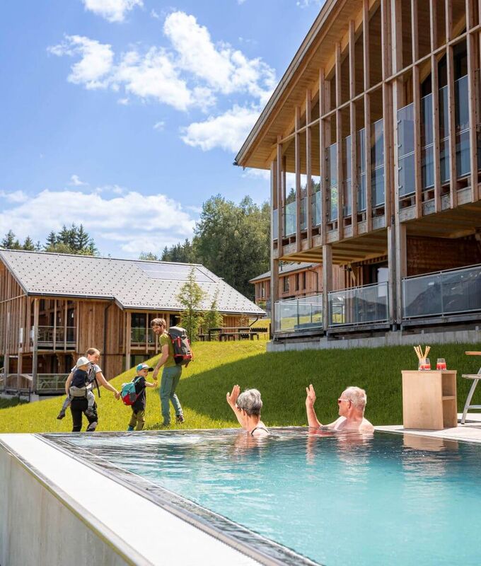 Großeltern winken der wandernden Jungfamilie aus dem privaten Pool ihres Ferienhauses zu.