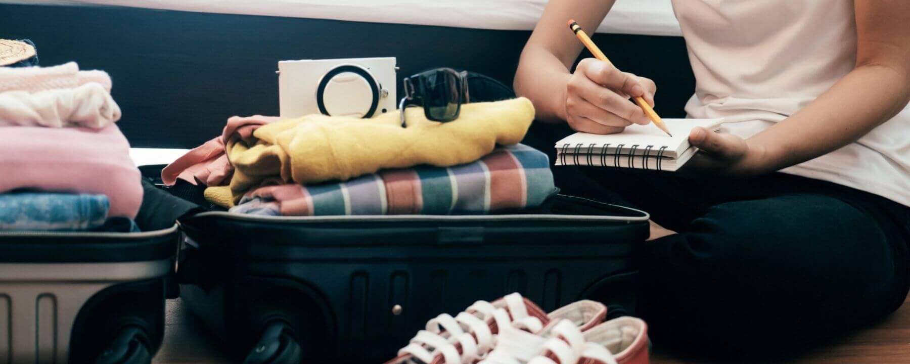 Young woman with a checklist sits on the floor in front of her open, fully packed suitcase.