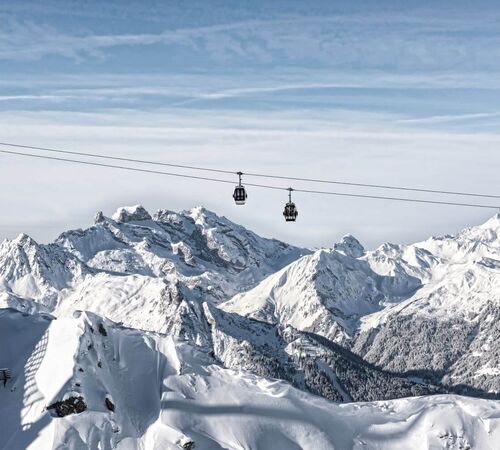Eine moderne Gondelbahn schwebt scheinbar oberhalb der verschneiten Gipfel im Hintergrund.