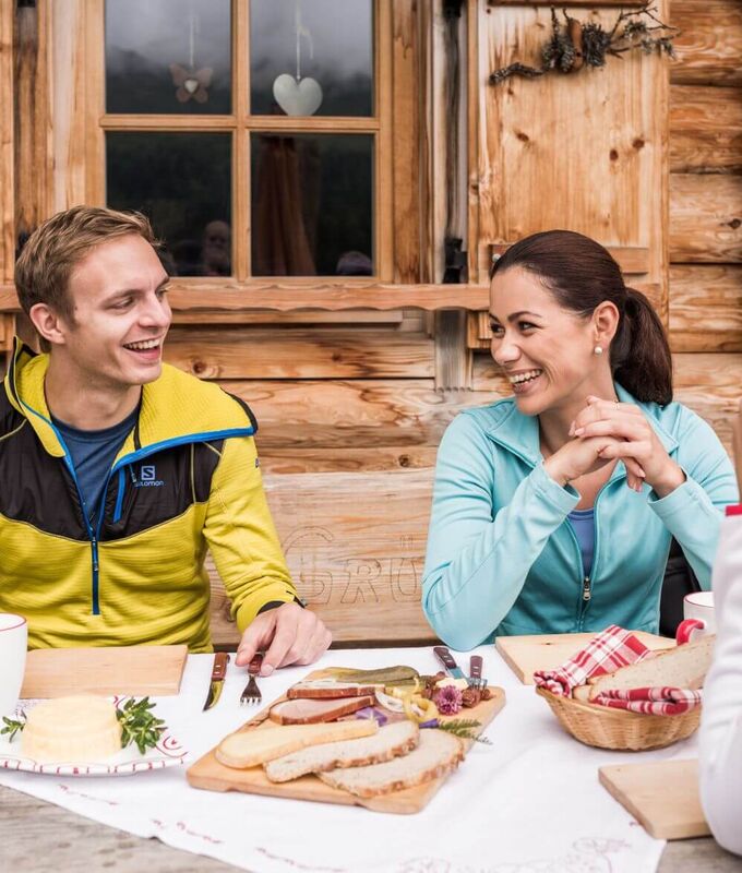Drei Menschen sitzen am Tisch einer Tiroler Alm und genießen regionale Produkte wie Brot und Speck