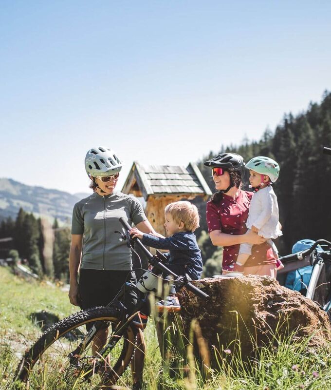 Zwei Mütter mit Fahrradhelmen und ihren Kindern machen Rast mit modernen E Bikes auf einer Almwiese inmitten einer alpinen Berglandschaft.