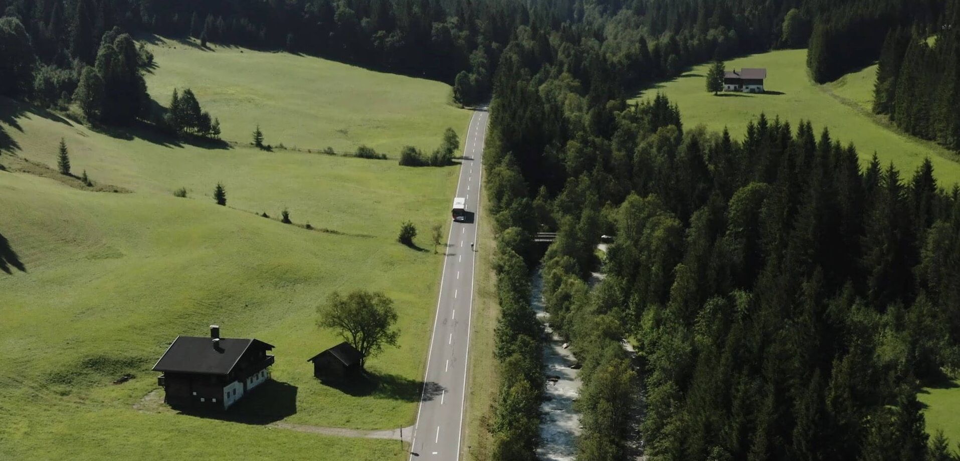 Bus fährt auf einer Landstraße umgeben von grünen Wiesen, Wald und Fluss.