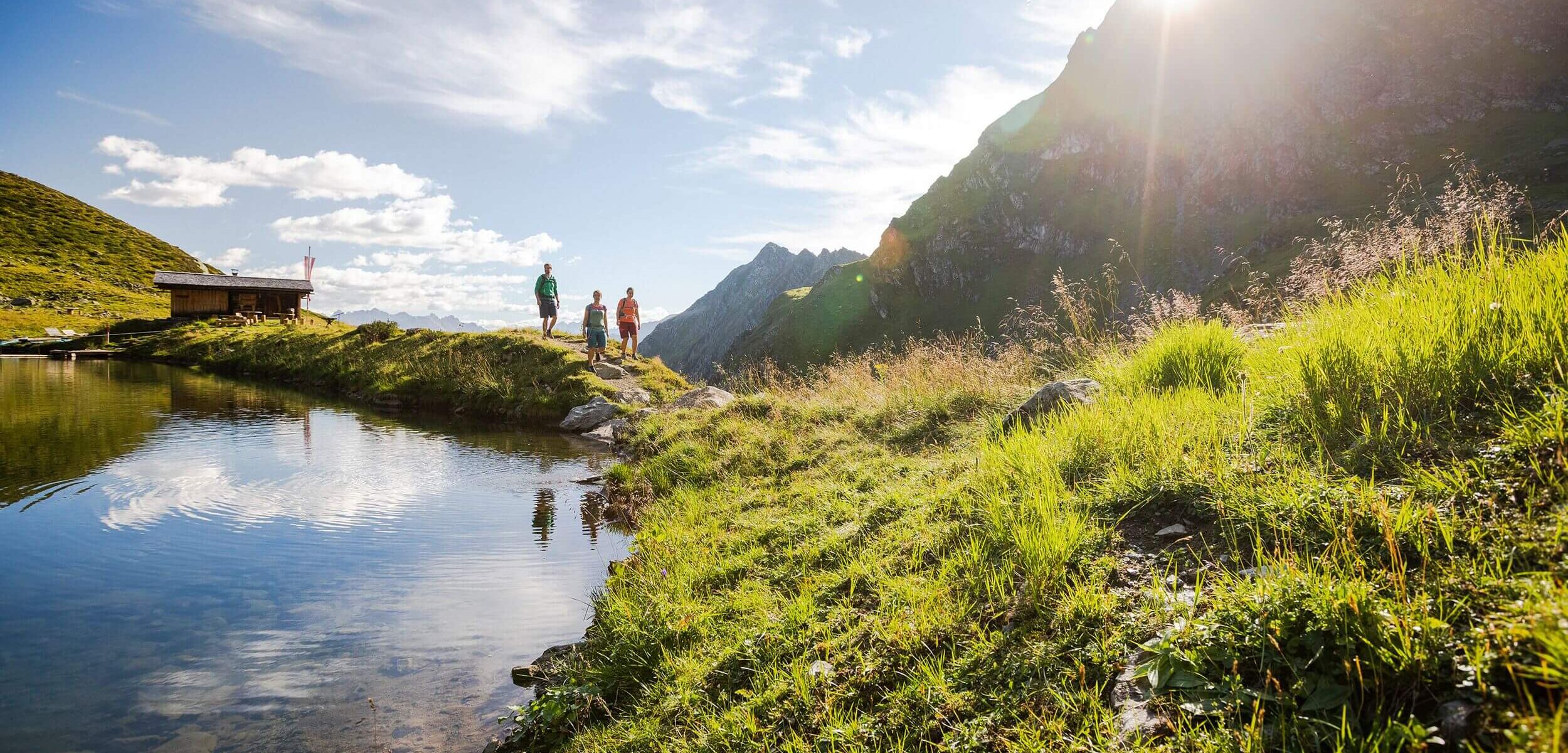 Familie beim Wandern entlang eines Bergsees mit traditioneller Tiroler Hütte und alpiner Bergkulisse im Hintergrund