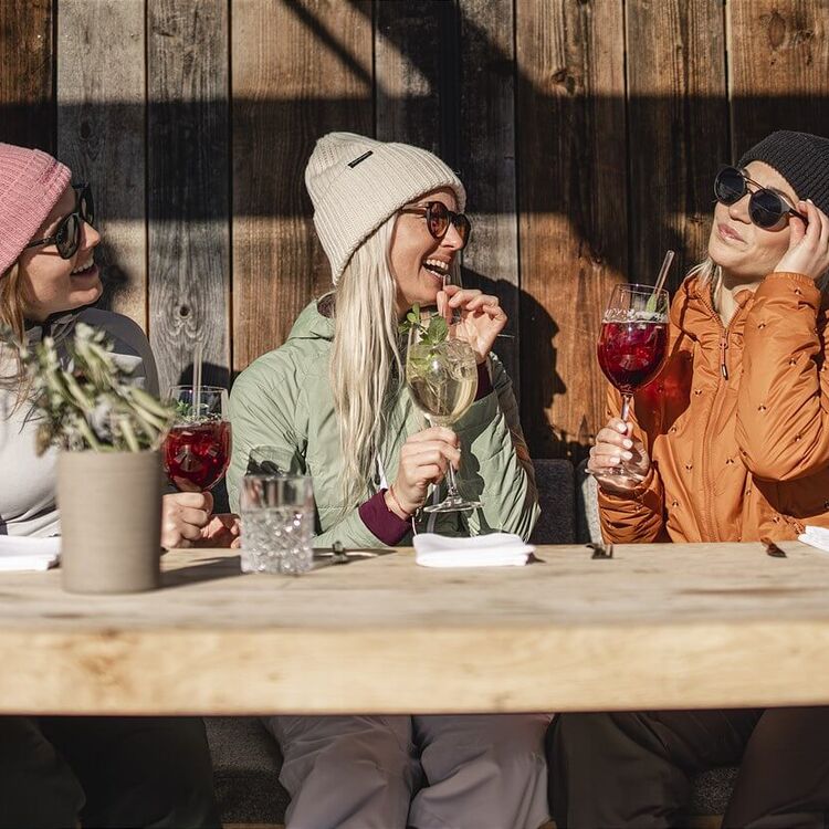 3 Freundinnen im Ski-Outfit sitzen vor einer Skihütte in der Sonne.