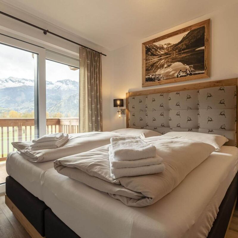 Helles Schlafzimmer mit bequemen Boxspring-Doppelbett, Holzelementen, gefalteten Handtüchern auf dem Bett, gepolstertem Kopfteil, Nachttischen und Balkon mit Aussicht auf herbstliche Natur und verschneite Berge