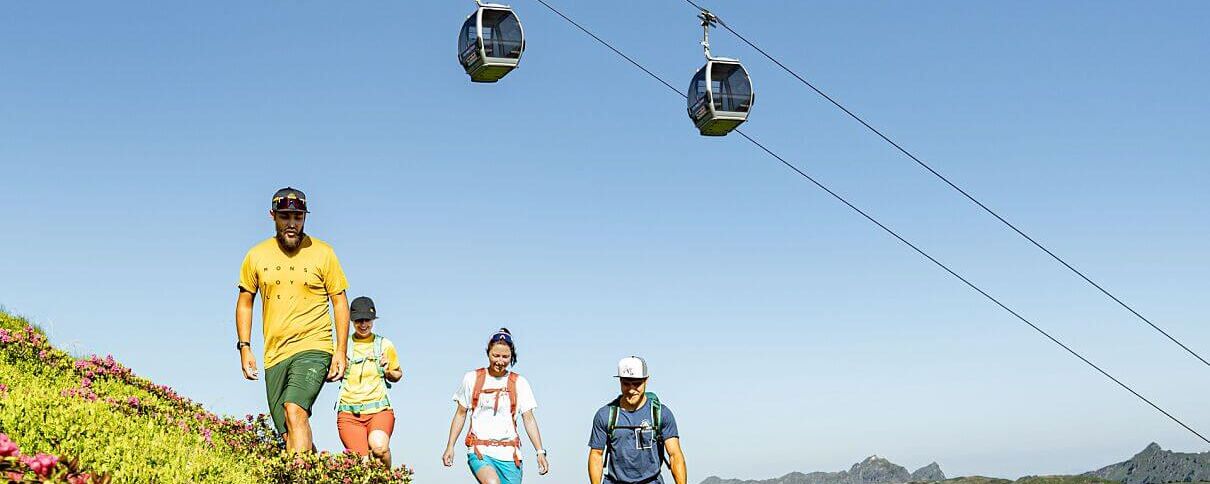 4 Freunde wandern unter einer Sommerbergbahn, im Vordergrund blühen die Almrosen.