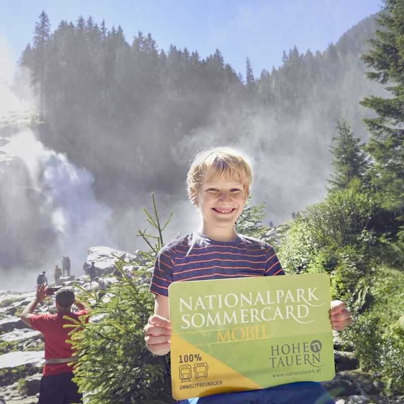 Junge steht mit der Nationalpark Sommercard in der Hand vor den Krimmler Wasserfällen.