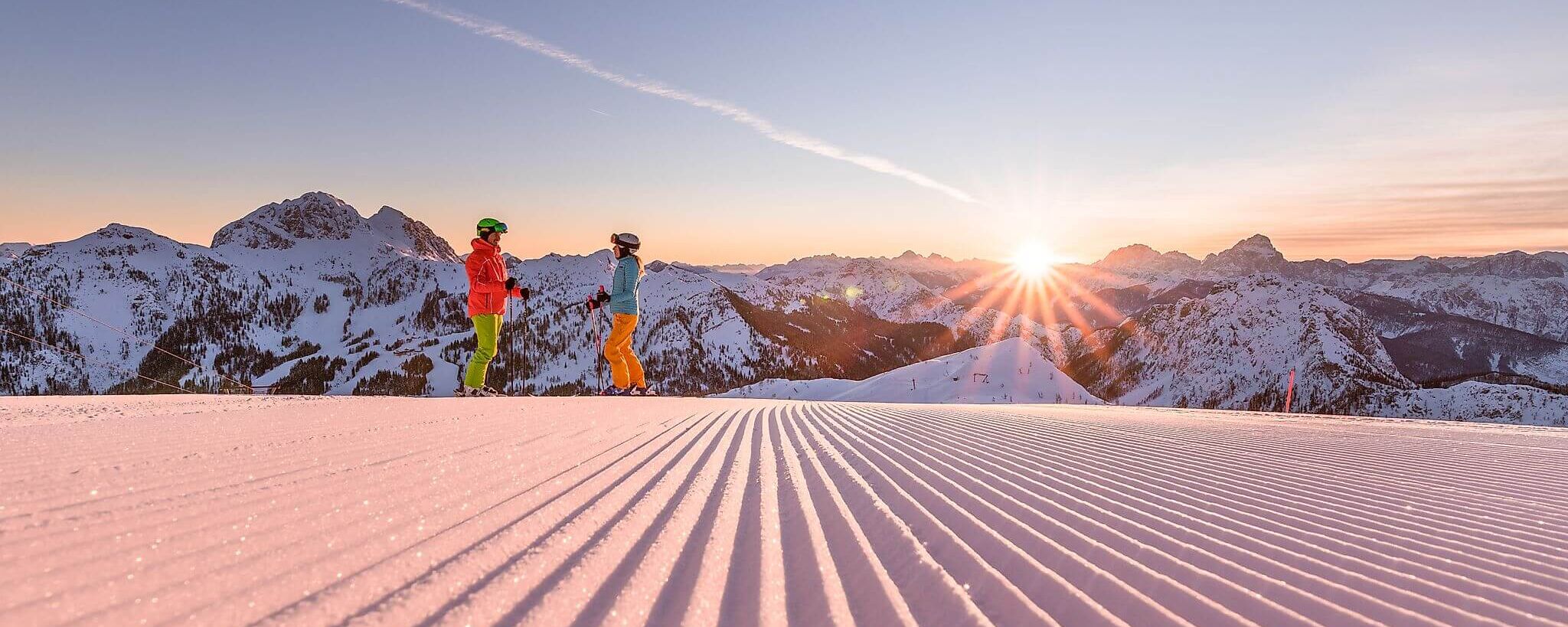 Zwei Skifahrer auf einer perfekt präparierten Piste im Skigebiet Nassfeld/Kärnten, Bergpanorama und Sonnenaufgang im Hintergrund.