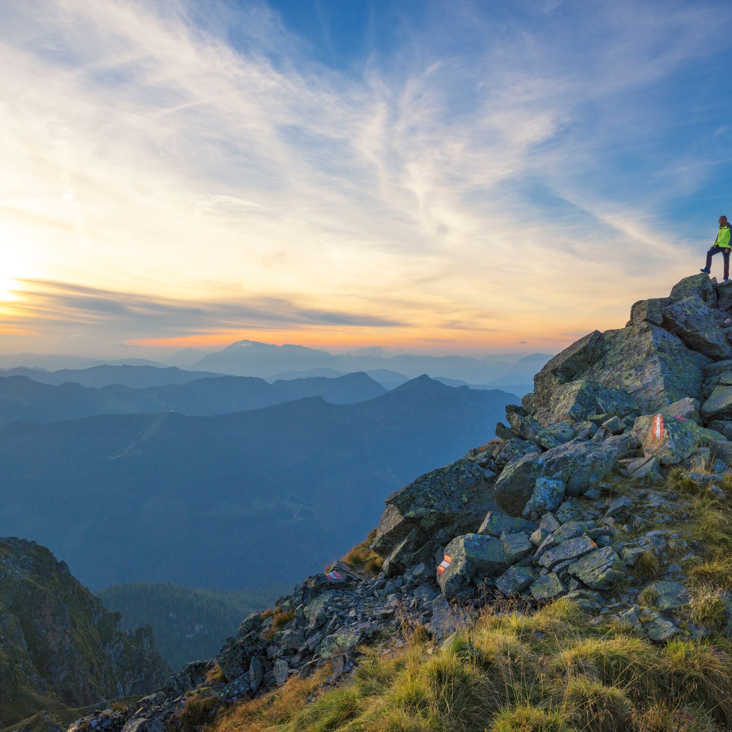 Zwei Wanderer stehen am Gipfelkreuz und blicken in die abendliche Berglandschaft mit weitem Panorama und warmem Licht