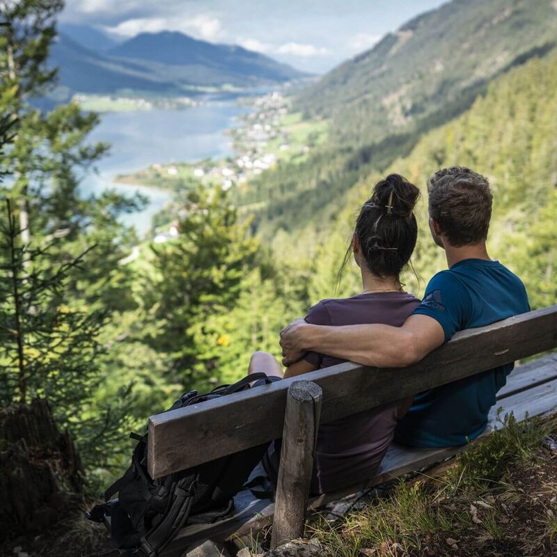 Ein Paar macht Rast auf einer Bank im Wald und genießt beim Wandern in Kärnten den Ausblick vom Berg auf einen See zwischen umliegenden Bergen