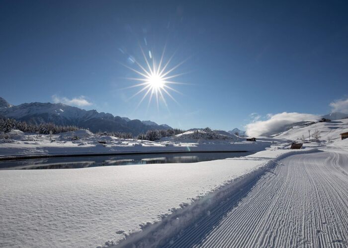 Winterlandschaft mit frisch gespurter Loipe, viel Schnee und sonnigem Ausblick auf einen klaren See