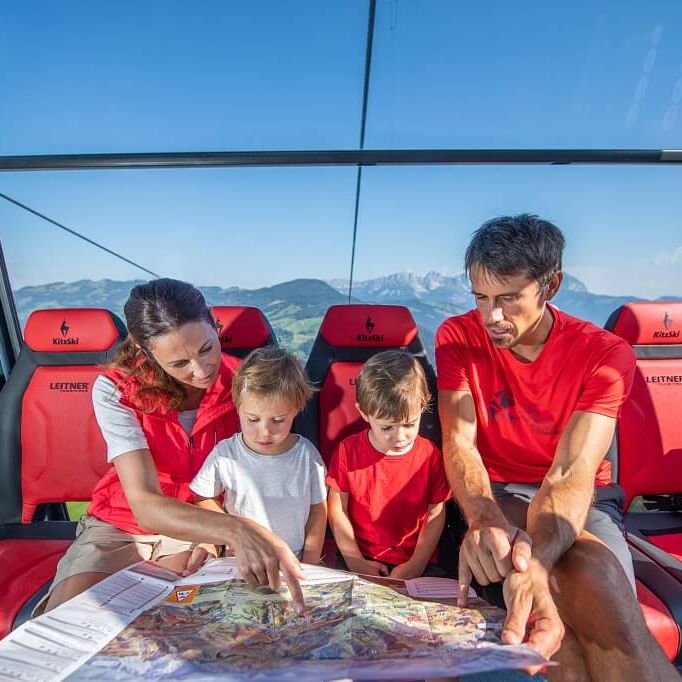 Familie mit 2 Kindern sitzt in einer verglasten Panoramagondel und studiert die Wanderkarte.