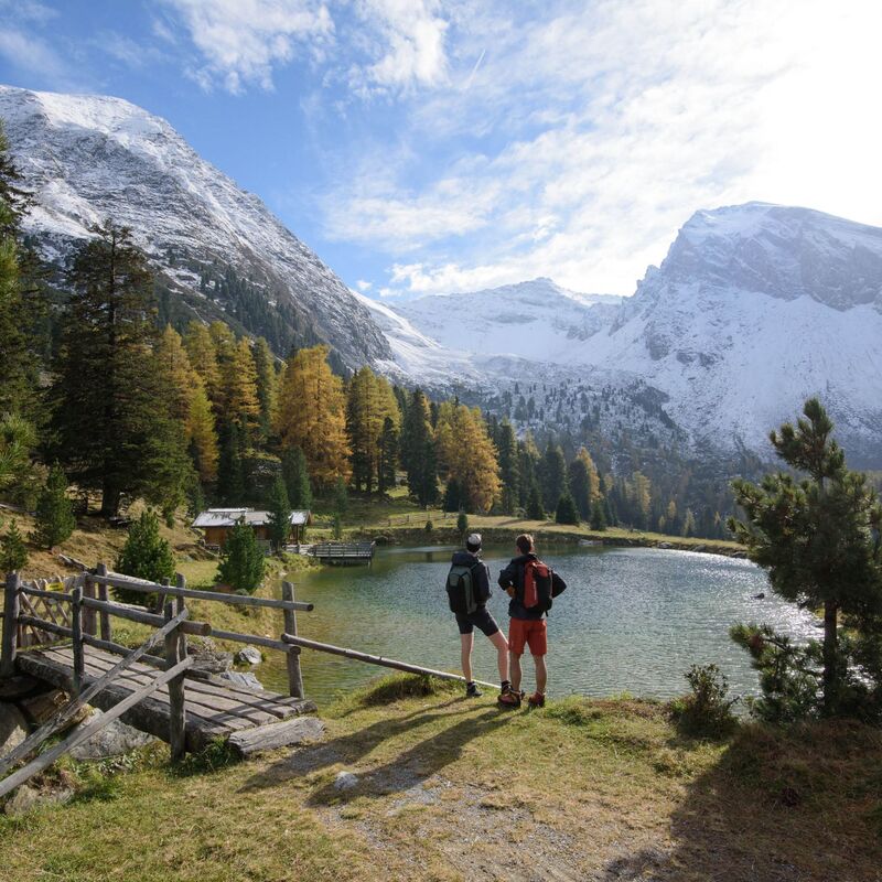 Wanderer stehen am klaren Bergsee im Zillertal, umgeben von herbstlichen Wäldern und leicht verschneiten Gipfeln