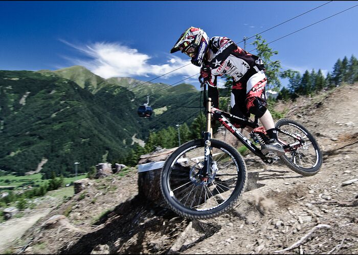 Ein Downhill-Biker in voller Schutzausrüstung fährt einen actionreichen Trail in alpiner Umgebung hinab.