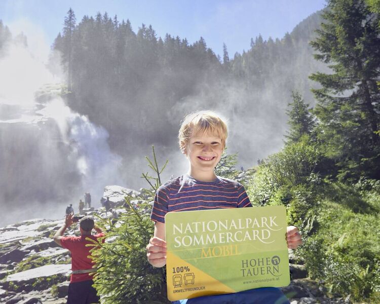 Junge steht mit der Nationalpark Sommercard in der Hand vor den Krimmler Wasserfällen.