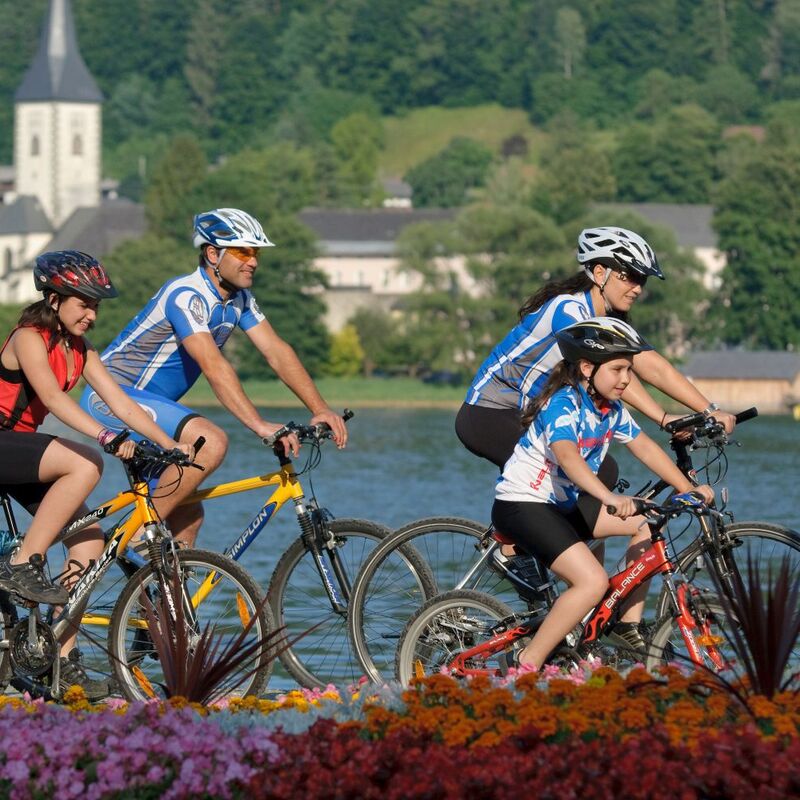 Eine Familie fährt gemütlich mit dem Fahrrad an einem See entlang und genießt die umliegende Berglandschaft in Österreich