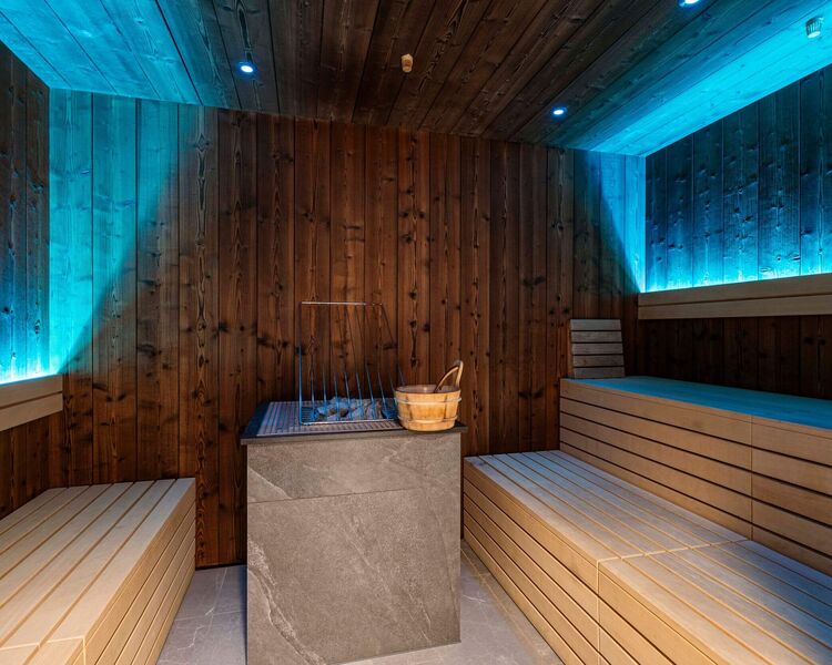 Sauna mit gemütlichen Holzbänken, Saunaofen aus Stein und entspannender Beleuchtung by ALPS RESORTS