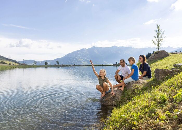 Familie entspannt am Wolfsee, Kinder spielen mit Wasser,umgeben von grüner Natur und Blick auf die Berge.