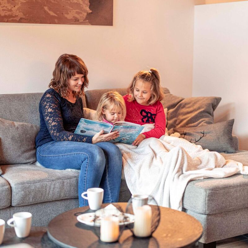 Mama liest mit Kindern Buch auf der Couch und alle genießen eine heiße Schokolade