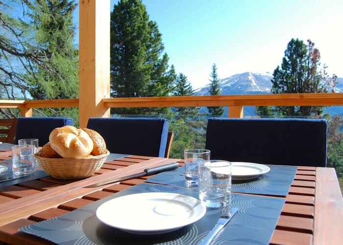 Frisch gedeckter Frühstückstisch auf einer Sonnenterrasse mit Blick auf grüne Berghänge und schneebedeckte Gipfel der Alpen.