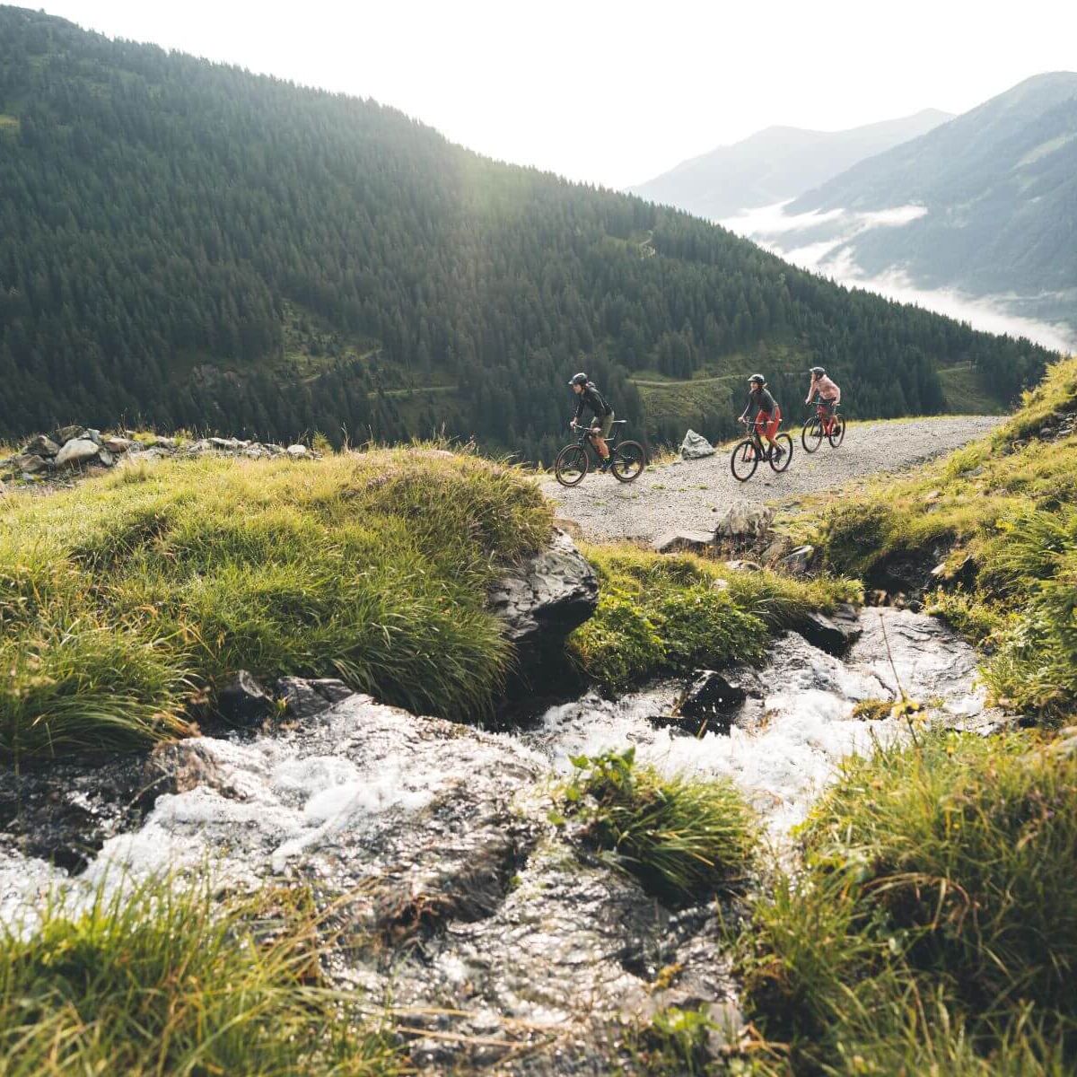 Drei Mountainbiker fahren auf einem breiten Bergweg entlang eines Gebirgsbachs mit Blick auf bewaldete Alpenlandschaft