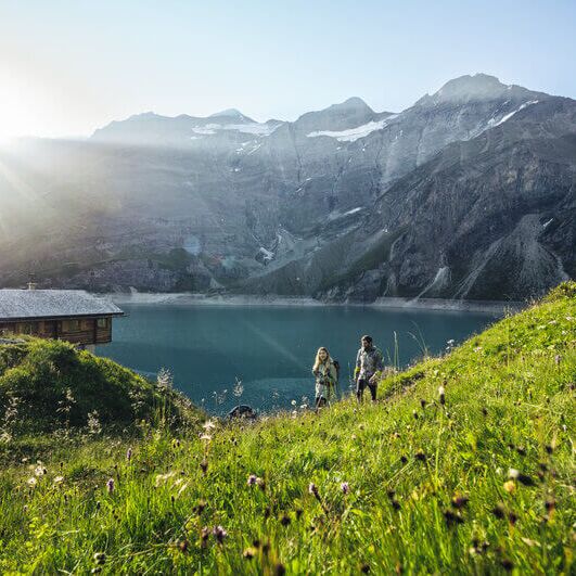 Zwei Wanderer spazieren an einer urigen Berghütte vorbei, entlang einer blühenden Almwiese mit Blick auf einen türkisfarbenen Stausee und die umliegende Bergkulisse im Salzburger Land.