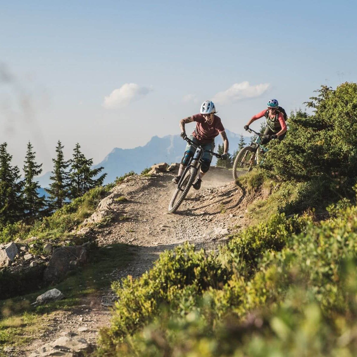 Zwei Downhill-Biker fahren actionreich eine alpine Strecke hinab, umgeben von beeindruckender Berglandschaft